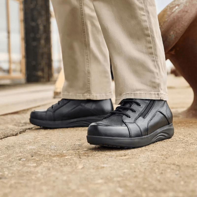 Mies yllään beiget housut ja mustat New Feet miesten varsikengät seisoo harmaalla pinnalla