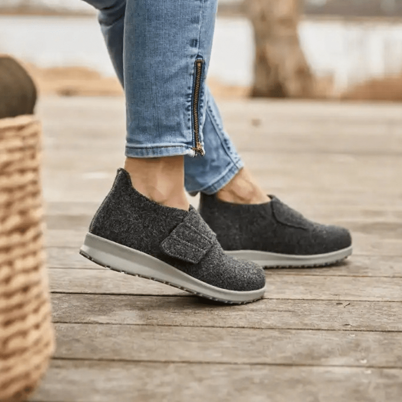 Nainen yllään antrasiitinharmaat New Feet tossut puulattialla, sivukuva keskittyen jalkoihin