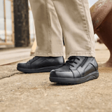 Mies yllään beiget housut ja mustat New Feet miesten varsikengät seisoo harmaalla pinnalla